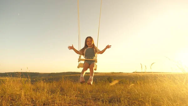 Joyeux enfant vole sur une balançoire dans le parc. Une petite fille joyeuse rit, rêve de devenir pilote et vole comme un avion. Maman secoue sa fille en bonne santé en se balançant sous un arbre au soleil. Enfance et famille. — Photo