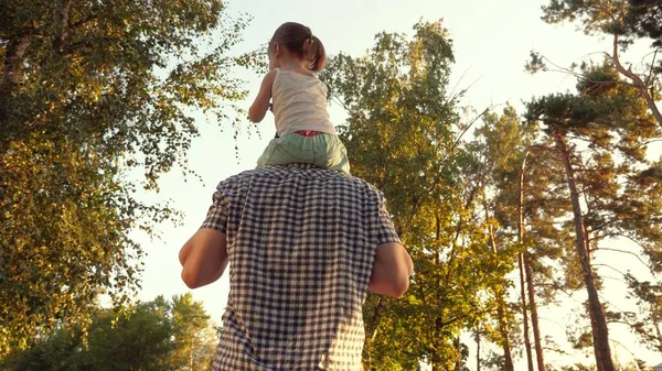 Papa porte les épaules de son enfant bien-aimé, dans le parc. Père marche avec sa fille sur ses épaules sous les arbres. enfant avec ses parents marche le jour de congé. Famille heureuse se relaxe dans le parc. — Photo