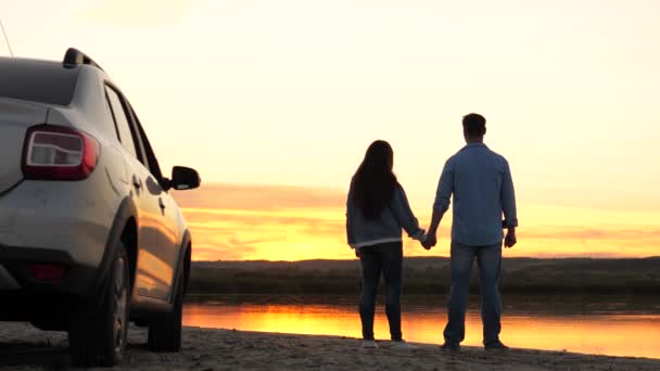 Tourists next to car, admiring sunrise, lake. Happy loving couple of travelers stand next to car and admire beautiful sunset over river on the beach. Free travelers, tourists. Family travel by car. — Stock Video