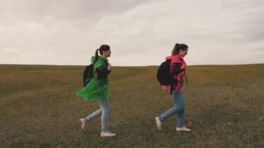 Happy women travelers walk hill, travelers teamwork. The concept of adventure and travel. Healthy girl tourists travel with backpacks in colorful raincoats in green and red. Hiking, outdoor adventure