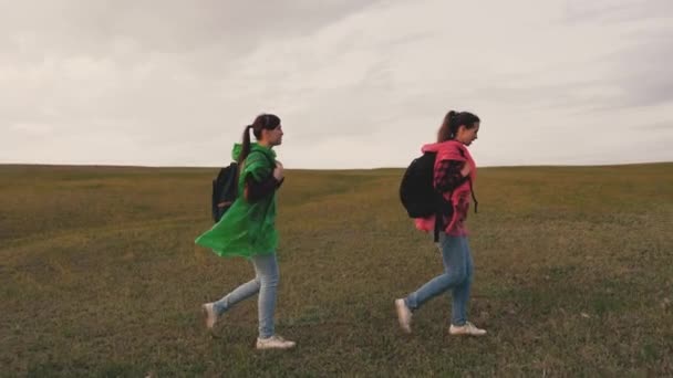 Happy women travelers walk hill, travelers teamwork. The concept of adventure and travel. Healthy girl tourists travel with backpacks in colorful raincoats in green and red. Hiking, outdoor adventure — Video Stock