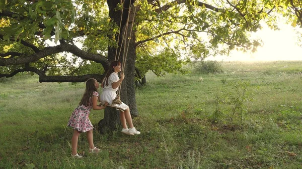 Las adolescentes disfrutan de volar en columpio en una noche de verano en el bosque. Niños balanceándose en un columpio en una rama de roble. Sueños de volar. Concepto de infancia feliz. Hermosas chicas están jugando en el parque. — Foto de Stock