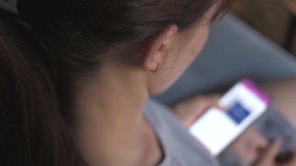 Mujer en casa sentada en el sofá en la sala de estar utilizando las compras de teléfonos inteligentes en la tienda en línea. mujer mira las mercancías en una tienda de ropa en línea. Compra ropa de moda directamente en tu smartphone. — Vídeo de stock