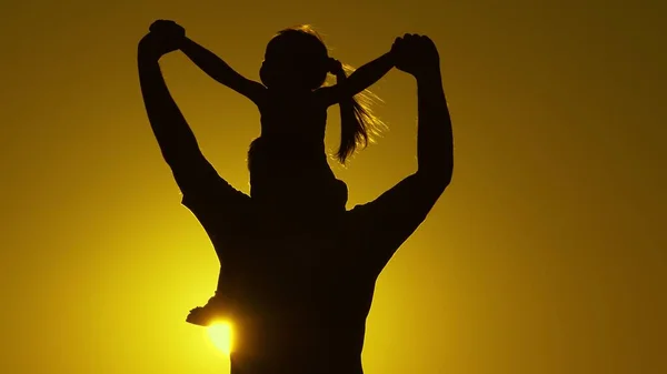 La famille heureuse se repose dans le parc. Père marche avec sa fille sur ses épaules au coucher du soleil. Silhouette d'un père porte sur les épaules de son enfant bien-aimé dans les rayons du soleil. Enfants en bonne santé marche avec papa. — Photo