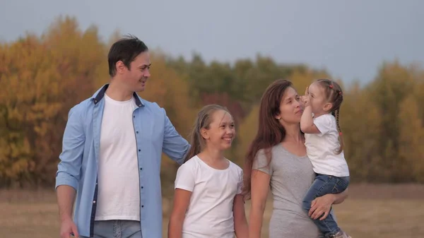 Gelukkige glimlachende familie die elkaars hand vasthoudt terwijl ze in het veld loopt, kleine dochter die in moeders armen rijdt. Gelukkige kinderen en ouders wandelen in het park, reizen en ontspannen. Familie speelt in de natuur met dochters. — Stockfoto