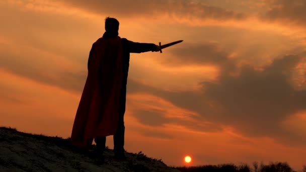 Juego de la legión romana en brillantes rayos de sol contra el cielo. Guerrero en una capa roja está parado en una montaña y agita su espada en la luz del atardecer. Caballero libre. Hombre libre juega superhéroe. — Vídeos de Stock