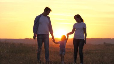 Mutlu aile parkta yürüyor. Güneşin altında el ele tutuşuyorlar. Anne, baba ve çocuk gün batımında mutlu, kız anne ve babasını elleriyle tutuyor. Sağlıklı çocuk atlıyor, ailesiyle oynuyor. Mutlu aile