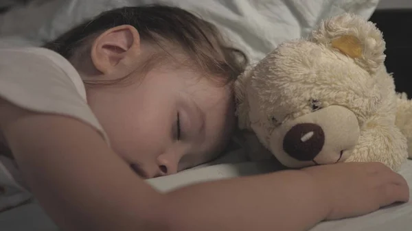Ребенок спит дома на диване в детской комнате. Спящий ребенок счастлив и беззаботен в постели, обнимая игрушку плюшевого медведя. Мать укрыла своего ребенка одеялом. Счастье во сне. — стоковое фото