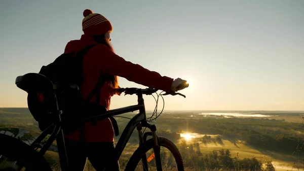 Happy cyclist woman resting in park and admiring beautiful landscape from mountain. Hiker healthy young woman stands on hill next to a bicycle, enjoying nature and sun. Free girl travels with bicycle