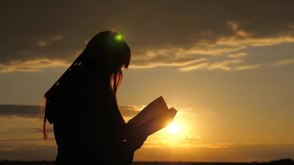 Una mujer lee un libro en rayos del sol. Chica pasa páginas de libro en rayos de atardecer, lee la Biblia al aire libre. Estudiando la Palabra de Dios al amanecer en la cima de la montaña. Buscando la verdad en las escrituras —  Fotos de Stock