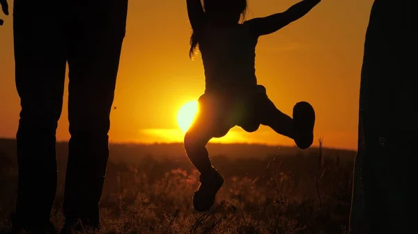 Il bambino sta saltando, tenendo le mani di papà e mamma al parco al sole. Una famiglia felice. La figlioletta gioca con papà e mamma sul campo al tramonto. Passeggiata con un bambino piccolo nella natura. Infanzia sana — Foto Stock