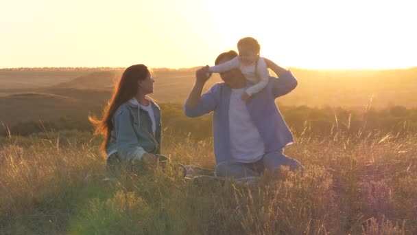 Papai bebê e mãe estão brincando ao ar livre. Família feliz brinca com sua filhinha no parque na grama em raios de pôr do sol. O miúdo sobe no pai. A filha abraça e ama seu pai e sua mãe. — Vídeo de Stock