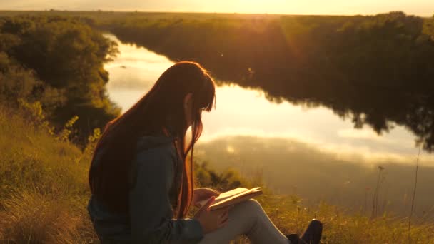 Viajante menina lê um livro em uma parada. Jovem estudante com livro didático ao ar livre. Uma jovem sentada em uma montanha em um gramado verde junto ao rio se preparando para um exame no parque. — Vídeo de Stock