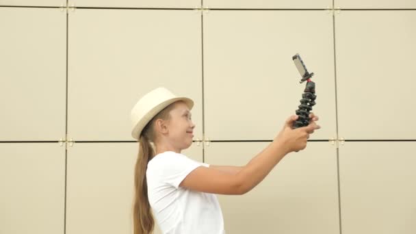 Ragazza alla moda in cappello fa selfie all'aperto. Un adolescente viene fotografato su smartphone utilizzando un piccolo treppiede. Free Girl blogger conduce una videoconferenza online da City Street. tecnologie digitali — Video Stock