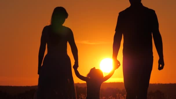 Щаслива сім'я, маленька донька стрибає, тримаючи за руки тата і маму в парку на сонці. Дитина грає з татом і мамою на полі у світлі заходу сонця. Прогулянка з маленькою дитиною в природі. дитинство, сім'я — стокове відео
