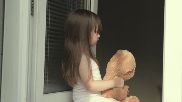 Beztroski dzieciak przytula swoją zabawkę. Dziecko na wiosnę bawi się zabawką przy otwartym oknie na parapecie. Mała dziewczynka w lecie bawi się z misiaczkiem, patrząc przez okno. Szczęśliwego dzieciństwa. Córka gra — Wideo stockowe