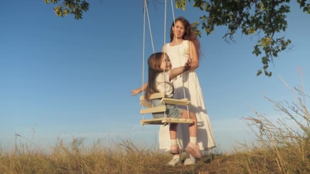 Feliz familia mamá sacude a un niño en un columpio y sonríe. El niño se eleva en vuelo con la madre. Un padre juega con su hija en un soleado día de verano. Trabajo en equipo. Diversión infantil al aire libre en el parque — Vídeos de Stock