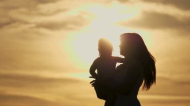 Mutlu bir aile, mutlu bir annenin silueti gün batımında uçan çocuğun etrafında dönüyor ve gülüyor. Anneliğin mutluluğu. Anneler Günü. Anne, kızlarını kucağında tutuyor gökyüzünün arka planına karşı