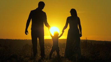 Küçük kız, güneşin arka planında anne ve babasının elini tutarak zıplıyor. Aile kavramı. Çocuk gün batımında anne ve babasıyla sahada oynuyor. Doğada küçük bir çocukla yürümek. çocukluk