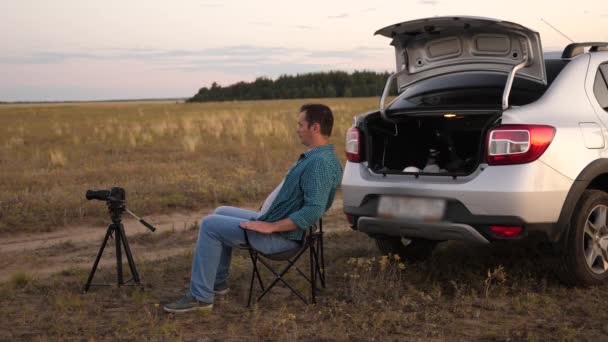 Un homme voyageur en voiture s'est arrêté à un camping le soir et prend des photos avec un appareil photo à côté de la voiture. Photographe freelance travaillant à l'extérieur, de beaux moments pour des photos et des vidéos. Le conducteur se repose. — Video