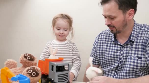 Zorgzame vader vrolijk helpen schattig kind om speelgoed vrachtwagen rijden, genieten van tgame. Gelukkige familie dochter en vader spelen speelgoed auto 's en pluche dieren van een haas en een beer. Ouder vader en kind spelen speelgoed thuis. — Stockvideo