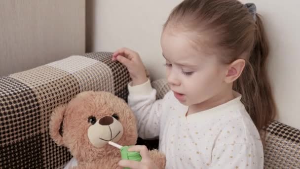 Barnflickan leker och behandlar leksaksbjörnen och helar sig själv. Lycklig familj. Grabben behandlar halsen, halsont med en spray mot halsont. Barn i pyjamas sjuk med influensa i rummet. Spray för hals — Stockvideo