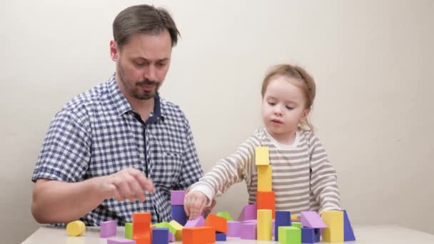Доглянутий батько і маленька дівчинка посміхаються, граючись з конструктором, щоб побудувати вежу і будинок з різнокольорових дерев'яних блоків. Щасливий батько допомагає милій дошкільній дочці грати в іграшкові кубики разом — стокове відео