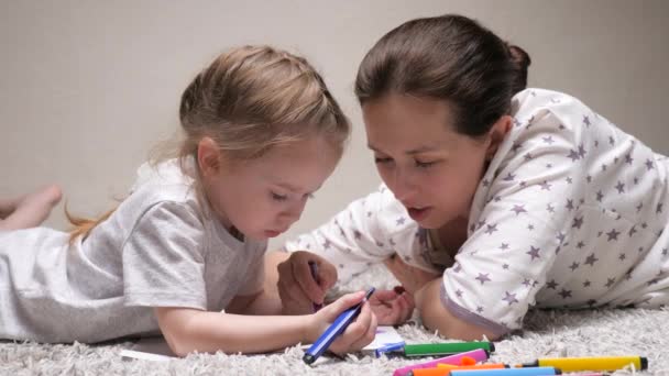 Mutlu aile evde yerde birlikte oynuyor. Anne, dadı, çocuk kıza resim yapmayı öğretiyor. Anne kızının kağıt üzerine resim çizmeyi öğrenmesine çok renkli kalemler ve keçeli kalemlerle yardım ediyor.. — Stok video