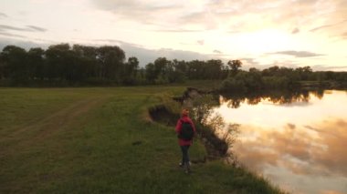 Sırt çantalı, gülümseyen ve el sallayan aktif, sağlıklı bir kadın beni takip ediyor. Parkta yürüyen gezgin, eğlence, biyoturizm, yürüyüş. Sırt çantasıyla nehir kenarında yürüyüş yapan genç bir kadın..