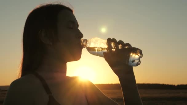 Young woman drinks water from a bottle after a workout. Jogging outdoors in rays of the sunset. Outdoor sports in the park in summer, spring, girl drinks clear cool mineral water after training. — Stock Video