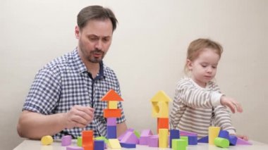 Baba ve küçük bir çocuk, bir kız, renkli ahşap küplerden bir ev ve kule inşa etmek için inşaat setiyle oynarken gülümsüyorlar. Mutlu ebeveyn babası tatlı okul kızına oyuncak blokları oynaması için yardım ediyor