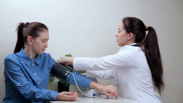 Un cardiólogo examina a una estudiante usando un tonómetro médico. Doctora que mide la presión arterial alta a una mujer joven en un hospital. Concepto asistencial de cardiología, diagnóstico precoz de enfermedades — Vídeo de stock