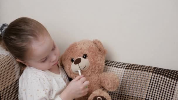 Traitez la gorge des enfants, le mal de gorge avec un spray de mal de gorge à lui-même et son jouet préféré à supporter. Enfant fille joue et traite l'ours jouet et se guérit. Enfant en pyjama malade de la grippe dans la chambre. — Video