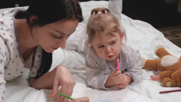 Мама, няня, учит девочку малювати. Щаслива сім'я бавиться вдома на ліжку. Мама допомагає дочці навчитися малювати на папері, розфарбовуючи багатокольорові олівці і ручки з чуттєвими кінчиками.. — стокове відео