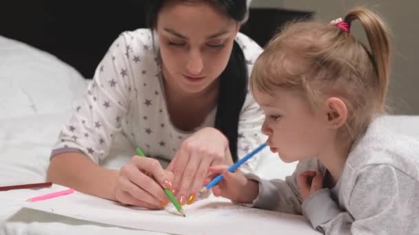 Madre, niñera, enseña a dibujar a la niña. Familia feliz jugando juntos en casa en la cama. Mamá ayuda a su hija a aprender a dibujar sobre papel coloreando con lápices multicolores y rotuladores. — Vídeo de stock