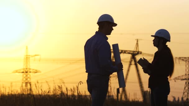 Gli ingegneri stanno discutendo il piano di lavoro. Due ingegneri in piedi sul campo con le torri elettriche al tramonto. La sagoma di ingegneri guarda la costruzione di potere ad alta tensione. Gruppo di ingegneri — Video Stock