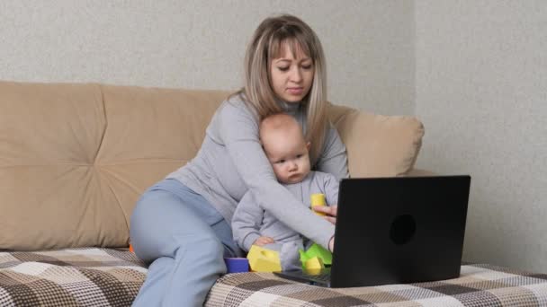 仕事中の母親はソファで赤ん坊と遊ぶ。赤ちゃんとノートパソコンで働く忙しい女性。居心地の良い家で彼女の腕の中で美しい赤ちゃんを持つ母親。女性のフリーランスの仕事。現代の母親 — ストック動画