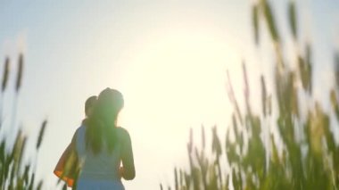 Anne ve küçük kızı yeşil buğday tarlasında yürüyor, sarılıyor ve öpüşüyorlar. Bir çocuk ve anne buğday tarlasında yürüyorlar. İyi yolculuklar. Bebek annesinin kollarında. Çiftçi kadın ve çocuk tarlada