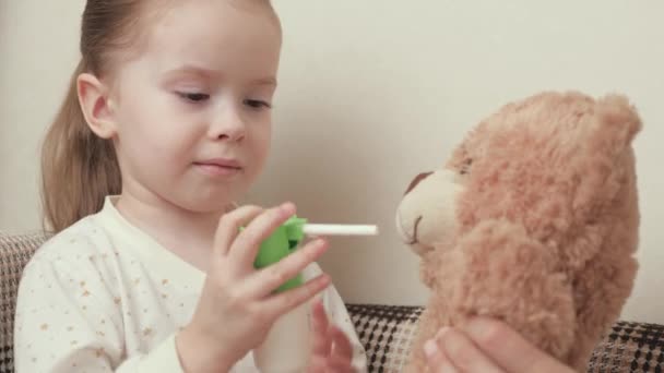 Barn behandle hals, ondt i halsen med en spray fra ondt i halsen til sig selv og hans foretrukne legetøj til at bære. Baby, pige leger og behandler legetøjsbjørn og helbreder sig selv. Dreng i pyjamas syg med influenza i rummet. – Stock-video