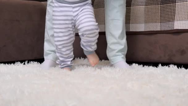 Bonne maman tenant la main aide mignon petit bébé faire les premiers pas à la maison. Le concept d'enfance et d'une famille heureuse. Bébé pieds nus fille garçon apprend à marcher tout en se tenant debout sur un sol chaud. — Video