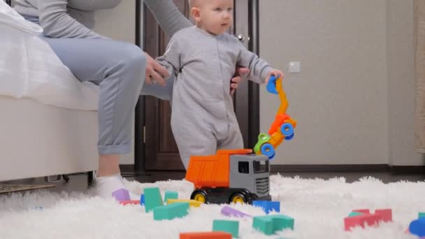 Ребенок играет дома с игрушечными машинами под присмотром своей матери. счастливая молодая няня играет с симпатичным малышом, ребенок весело бегает по ковру дома. счастливая семья. образовательные игры — стоковое видео