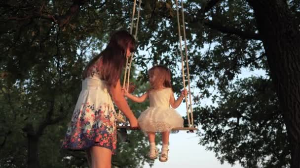 Mom plays with her child daughter, they swing on swing in park on playground under tree. A healthy family is playing in park. A mother shakes her healthy little daughter on a swing under a tree in sun — стоковое видео