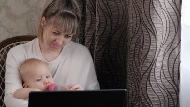 The mother works at computer with the child. The kid sits with his mother, drinks, eats from a baby bottle. Freelance woman with infant. Business woman working at home on laptop with toddler at table — Stock Video