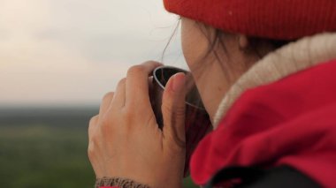 Tourist is drinking tea from a mug in sun. Traveler Woman holds metal mug with hot coffee in her hands and looks at sunrise from mountain. Free girl hiker admiring landscape. Travel, camping