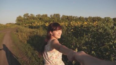 Free girl and guy hand in hand, walk on field with sunflowers at sunset, hold hands. Young woman travels through countryside with her boyfriend, they run across field of a blooming sunflower.