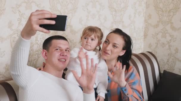 Happy young caucasian family with baby boy waving to friends while talking during smartphone video call at home. Mother, father and daughter bloggers in room communicate using a modern gadget online. — ストック動画
