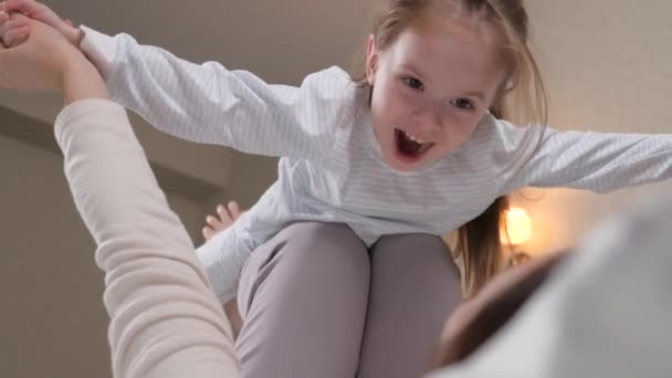 Bir çocuk, bir kız, oynuyor, gülüyor ve annesinin kollarında uçuyor. Bir çocuk ve genç bir anne evdeki kanepede birlikte uçak oynuyorlar, uçmayı hayal ediyorlar. Mutlu bir aile. Aile jimnastik yapıyor. — Stok video