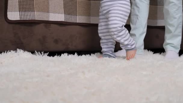 Bonne maman tenant la main à l'enfant aidant mignon petit bambin à faire les premiers pas à la maison, jeune garçon tenant canapé. Famille, enfance. Bébé pieds nus fille garçon apprend à marcher tout en se tenant debout sur le sol chaud — Video