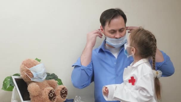 Ο πατέρας, ο γιατρός, διδάσκει την κόρη του να φοράει ιατρική μάσκα. Χαριτωμένο παιδάκι, κόρη, ντυμένη με λευκό παλτό γιατρού, παίζει με τον μπαμπά γιατρό. Αστείο κορίτσι παίζει ένα παιχνίδι με τον μπαμπά γιατρό και ασθενή — Αρχείο Βίντεο