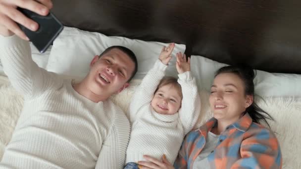 Feliz familia joven con una niña, son fotografiados en un teléfono inteligente en la cama. La madre, el padre y la hija blogueros en la habitación se comunican usando un gadget moderno en línea. Familia joven moderna — Vídeos de Stock